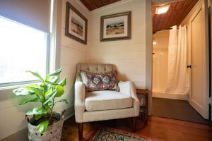 A cozy corner with a beige armchair and patterned cushion, next to a potted plant by a window. Two framed pictures hang on the wall, and a bathroom with a white shower curtain is visible in the background.