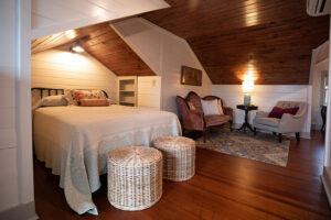 Cozy attic bedroom with a sloped wooden ceiling, a double bed, two wicker stools, two armchairs, a small table with a lamp, and a patterned rug on polished wooden floors.