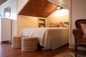 Cozy bedroom with a sloped wooden ceiling, a large bed with white bedding, two wicker stools at the foot of the bed, a white dresser, and an upholstered armchair on hardwood floors.