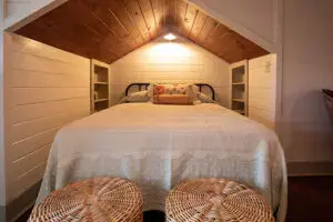 A cozy loft bedroom with a sloped wooden ceiling, a neatly made bed with decorative pillows, open shelving on each side, and two woven stools at the foot of the bed. Warm lighting creates a welcoming atmosphere.