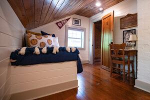 Cozy attic bedroom with a slanted wooden ceiling, a bed with blue and white bedding, decorative pillows, a small window, a wooden desk and chair, a lamp, and wall decorations including a pennant and framed pictures.