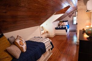 Cozy attic bedroom with wooden floors and sloped wood-paneled ceiling, featuring two beds with colorful pillows and striped blankets, a small window, and a dresser with a lamp and globe.