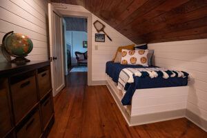 A cozy attic bedroom with wood-paneled ceiling, a bed with blue and white bedding, decorative pillows, a globe on a cabinet, and tennis rackets hung on the wall; an open door leads to another room.