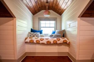 Cozy reading nook with a sloped wooden ceiling, white shiplap walls, a window, and a cushioned bench covered with a lion-patterned blanket and several decorative pillows.