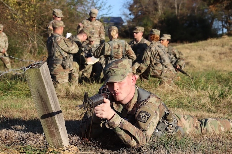 A soldier in camouflage uniform lies prone on the ground aiming a training rifle, while a group of soldiers gathers and writes notes in the background on a grassy field.