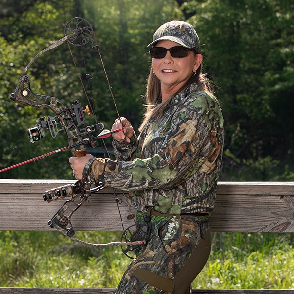 A woman in camo gear and sunglasses stands outdoors, holding a compound bow. She is smiling and standing next to a wooden railing, with greenery and trees in the background.