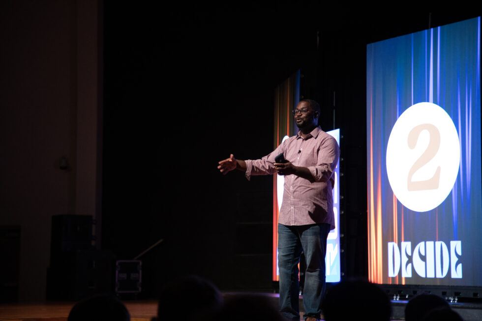 A man in a pink shirt and jeans stands on stage with arms open, speaking in front of a large screen displaying the number 2 and the word DECIDE in bold letters.