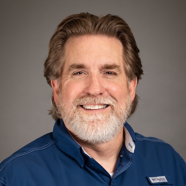 A smiling middle-aged man with short brown hair and a neatly trimmed gray beard, wearing a dark suit, white shirt, and blue striped tie, posed against a solid gray background.