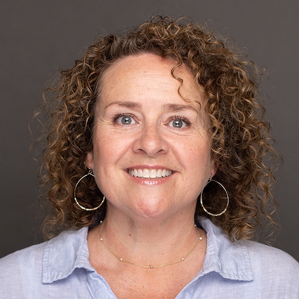 A woman with curly brown hair smiles at the camera. She is wearing large hoop earrings, a delicate necklace, and a light blue collared shirt, with a plain dark background behind her.