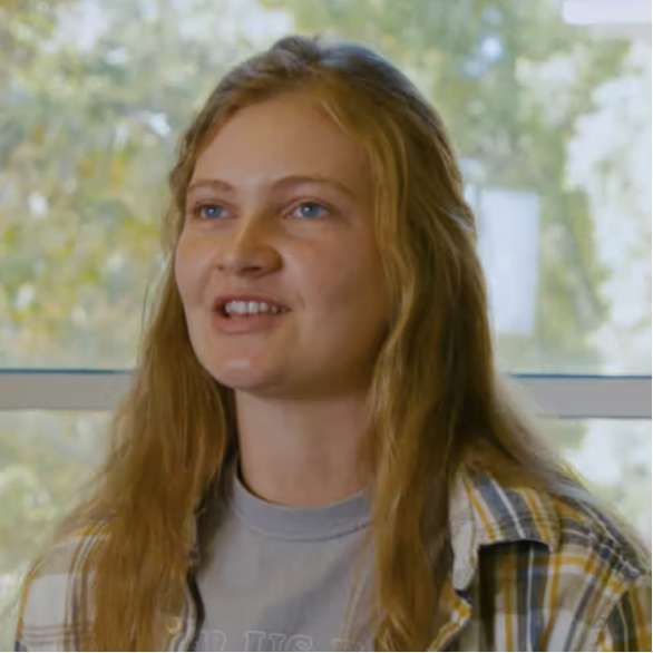 A young woman with long blonde hair, wearing a plaid shirt over a gray t-shirt, smiles while sitting indoors with a window and green trees in the background.