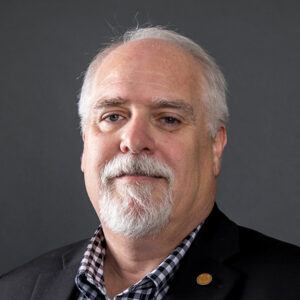 A middle-aged man with white hair and a beard, wearing a black checked shirt and black blazer, poses against a plain dark background.