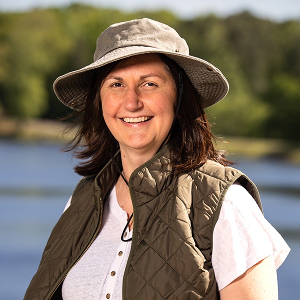 A woman wearing a wide-brimmed hat and a quilted vest smiles outdoors by a lake, with trees and blue sky in the background.