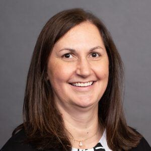 A woman with straight brown hair, wearing a black jacket over a black-and-white top, smiles at the camera in front of a plain gray background.