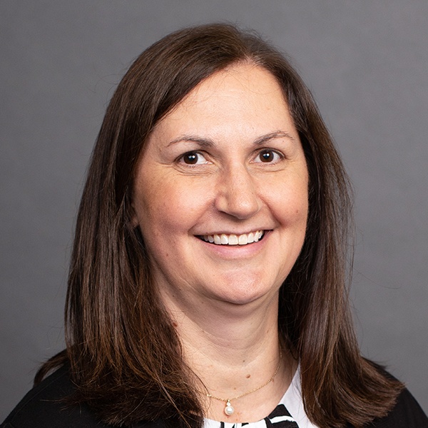 A woman with straight brown hair, wearing a black jacket over a black-and-white top, smiles at the camera in front of a plain gray background.