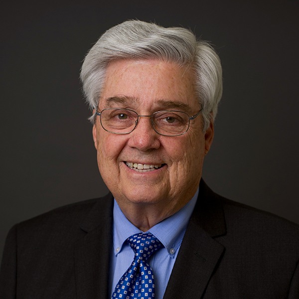An older man with gray hair and glasses smiles at the camera. He is wearing a dark suit, blue dress shirt, and a blue patterned tie, with a plain dark background behind him.