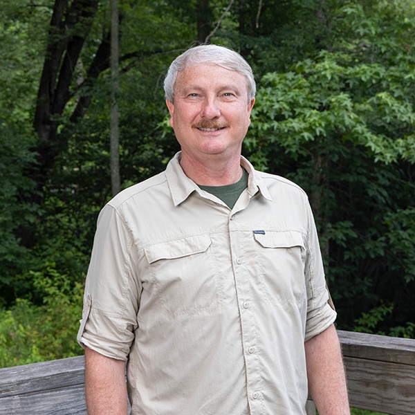 A middle-aged man with gray hair and a mustache stands outdoors on a wooden deck, smiling, with green trees and foliage in the background. He is wearing a light beige button-up shirt.