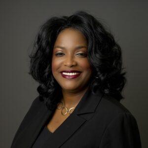 A woman with long, curly black hair smiles warmly. She is wearing a black blazer, a black top, and a gold necklace with circular links, posed against a plain dark background.