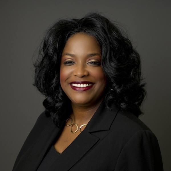 A woman with long, curly black hair smiles warmly. She is wearing a black blazer, a black top, and a gold necklace with circular links, posed against a plain dark background.