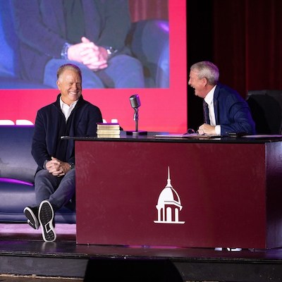 Two men sit and smile on a stage during an interview. One sits casually on a couch, and the other sits behind a desk with a microphone and books. A maroon backdrop features a white dome logo.
