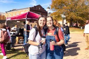 Two young women smile while holding drinks at an outdoor event. Behind them, people gather near a tent with refreshments. Autumn trees and a brick building are visible in the background under a sunny sky.