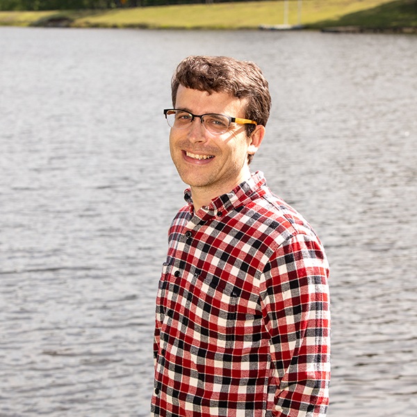 A man wearing glasses and a red plaid shirt stands smiling by a calm lake, with grassy hills in the background under a bright sky.