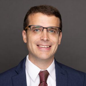 A man wearing glasses, a navy blue suit, white shirt, and red patterned tie, smiling against a plain dark gray background.