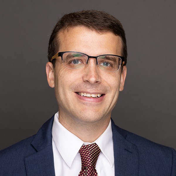 A man wearing glasses, a navy blue suit, white shirt, and red patterned tie, smiling against a plain dark gray background.