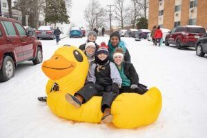 Five people sit and smile on a large inflatable yellow duck in the snow, surrounded by parked cars and buildings, with others walking in the background on a winter day.