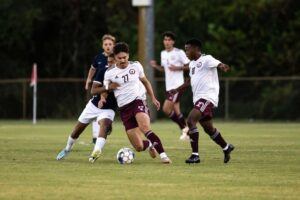 A soccer player in a white jersey with the number 21 dribbles the ball, pursued closely by opponents. Another teammate in white runs alongside on a grassy field, with trees and a fence in the background.