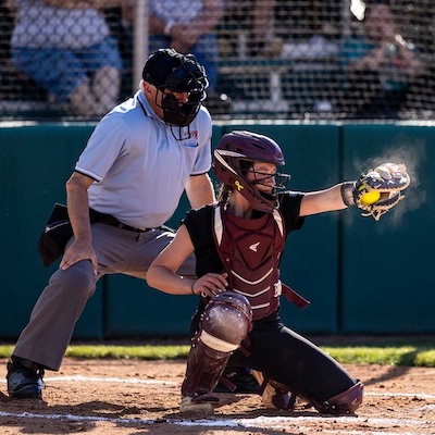 A softball catcher in full gear catches a yellow softball, with dust flying from the impact, as an umpire stands behind her during a game. Spectators watch from behind a fence in the background.