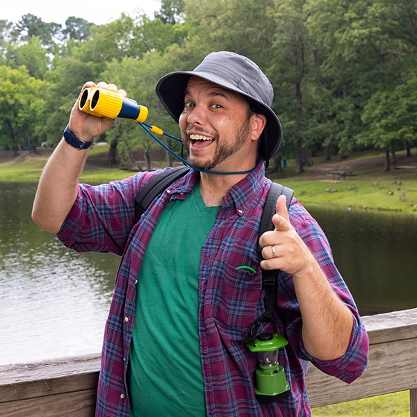 A smiling man in a hat and plaid shirt stands by a lake, holding yellow binoculars and pointing at the camera. He has a backpack and a green water bottle, with trees and water in the background.