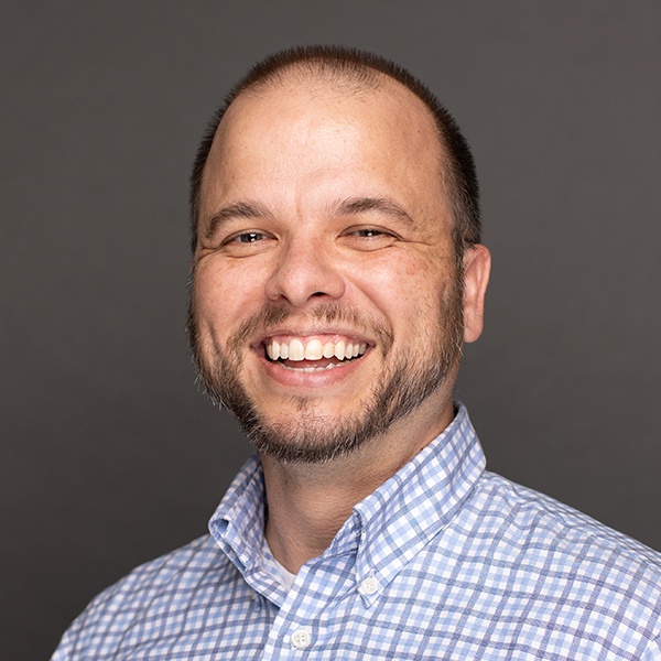 Smiling man with short brown hair and a trimmed beard, wearing a light blue and white checkered button-up shirt, standing in front of a plain gray background.