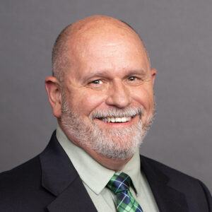 A smiling older man with a bald head, gray beard, and mustache, wearing a dark suit jacket, light green shirt, and green plaid tie, posed in front of a plain gray background.