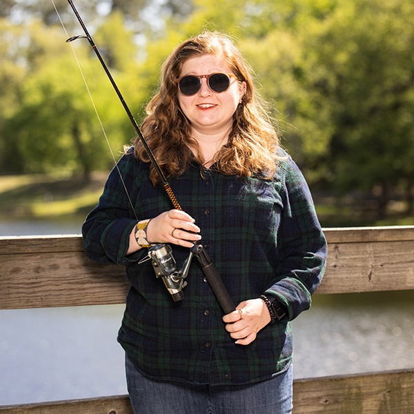 A woman with long, wavy hair wearing sunglasses and a plaid shirt stands on a wooden bridge, holding a fishing rod, with a pond and green trees in the background.
