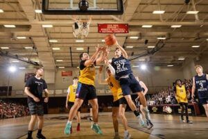 Two basketball teams compete in a gym; a player in a navy jersey jumps to shoot while a player in yellow blocks, with other players and spectators visible in the background.