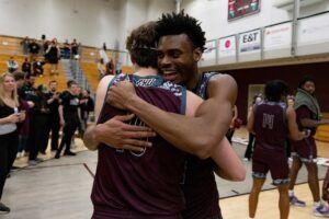 Two basketball players in maroon uniforms hug and smile on a gym court after a game, with teammates and spectators in the background celebrating.