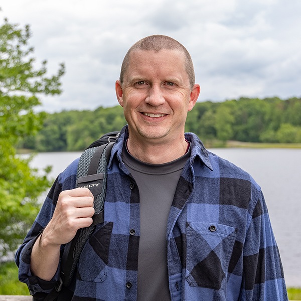A man with short hair, wearing a blue and black plaid shirt over a gray t-shirt, stands outdoors by a lake, smiling and holding a backpack strap, with trees and water in the background.