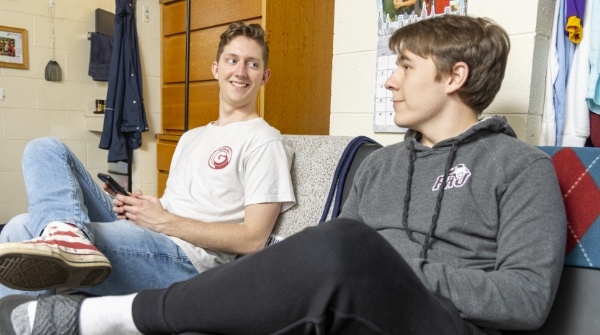 Two young men sit on a couch in a dorm room, smiling and talking. One holds a phone, while the other wears a hoodie. The room has clothes, a dresser, and various decorations in the background.