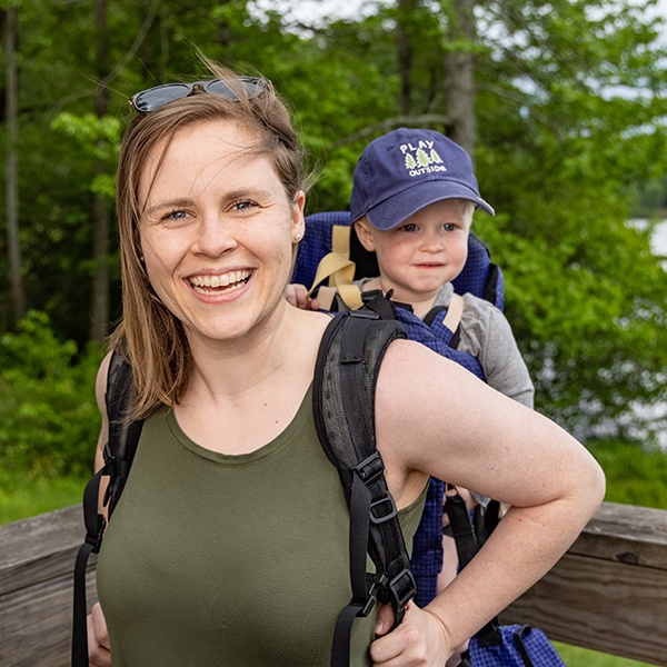 A smiling woman with shoulder-length brown hair carries a young child in a hiking backpack. The child wears a navy “Play Outside” cap. They are outdoors with green trees in the background.
