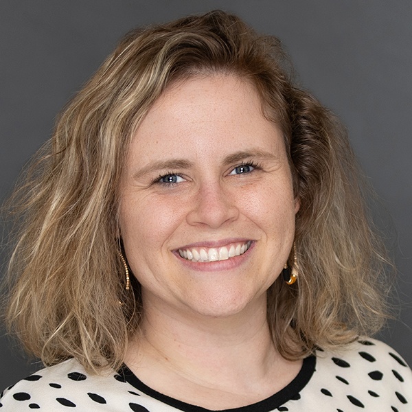 A woman with wavy blonde hair smiles at the camera. She is wearing a cream top with black polka dots, gold earrings, and is posed in front of a plain gray background.