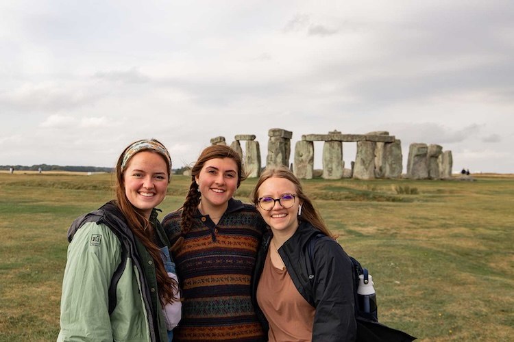 Three young women smiling and posing together in front of Stonehenge on a cloudy day, standing on green grass with the ancient stone structure visible in the background.