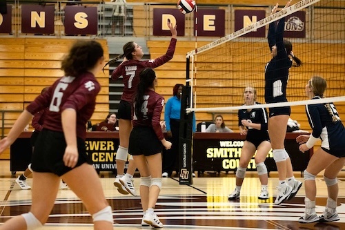 Four volleyball players in maroon jerseys face two players in navy jerseys who are attempting to block a spike at the net in a gymnasium with empty bleachers and banners in the background.