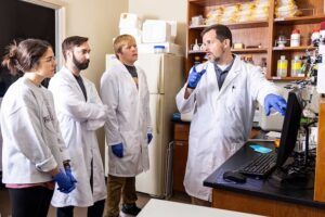 A scientist in a lab coat and gloves explains something to three students, also in lab coats and gloves, in a laboratory with shelves of bottles and scientific equipment.