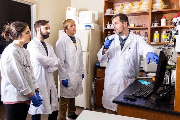 A scientist in a lab coat and gloves explains something to three students, also in lab coats and gloves, in a laboratory with shelves of bottles and scientific equipment.