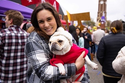 A smiling woman in a plaid coat holds a French bulldog wearing a red sweater and gray hat at an outdoor event with people and colorful booths in the background.