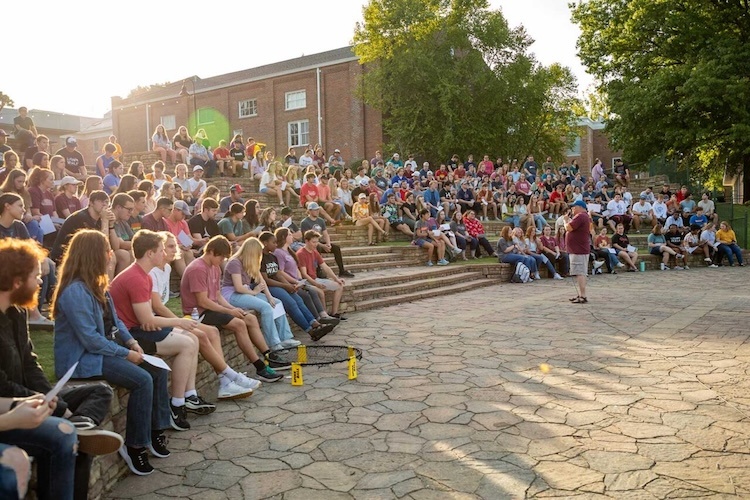 A large group of people sit on outdoor stone steps, listening to a person speaking in front of them on a sunny day. Trees and a brick building are visible in the background.