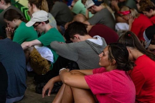 A group of young people sit closely together on the ground, many with heads bowed and eyes closed, appearing thoughtful or in prayer. Most are dressed casually in colorful t-shirts.