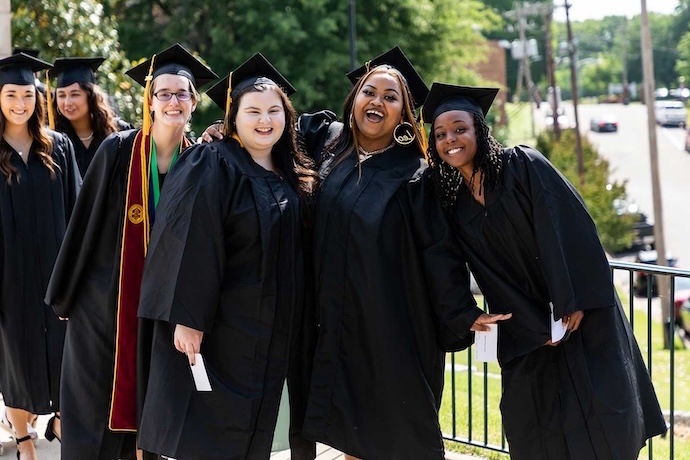 Five graduates in caps and gowns smile and pose together outdoors on a sunny day, celebrating their achievement. Some hold diplomas, and greenery and a street are visible in the background.