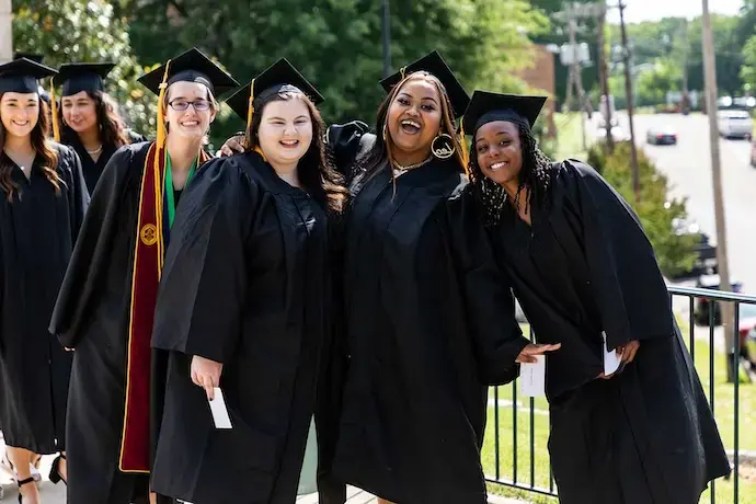 A group of students smiling in the commons in their graduation regalia