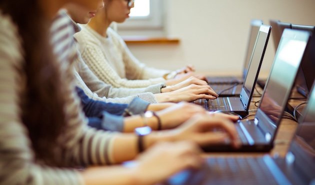 Several people sit in a row, typing on laptops at a desk. The image focuses on their hands and keyboards, with faces partly out of frame. They appear to be in a classroom or study environment.